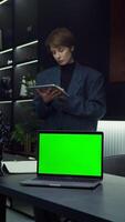 An open laptop with a green screen stands on the desk of a young businesswoman working on a tablet, standing behind, vertical . Chromakey, layout, copy space. in 4k format, red komodo. video