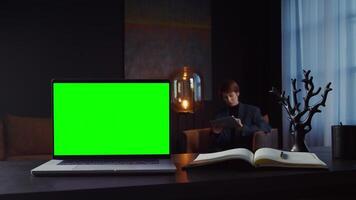 An open laptop with a green screen stands on the desktop of a young businesswoman using a tablet while sitting in a chair on the background, in a stylish interior. in 4k format, red komodo. video