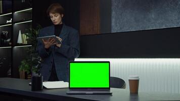 An open laptop with a green screen stands on the desk of a young businesswoman using a tablet with a serious look, standing behind. Chromakey, layout, copy space. in 4k format, red komodo. video