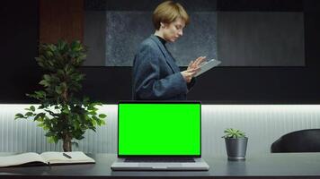 An open laptop with a green screen stands on a desktop in a stylish office of a young businesswoman using a tablet while walking past the table. Mockup, copy space. in 4k format, red komodo video