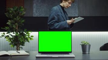 An open laptop with a green screen stands on a desktop in a stylish office of a young businesswoman using a tablet in the background. Mockup, chroma key, copy space. in 4k format, red komodo. video