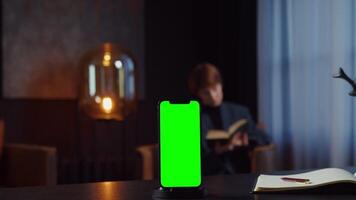 A mobile phone with a green screen stands on desktop of businesswoman reading a book in a chair in the background. focused on smartphone, chroma key, copy space. in 4k format, red komodo video