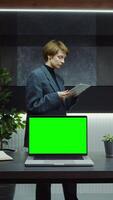 An open laptop with green screen stands on desktop in the office of young businesswoman using tablet while walking around the office. Mockup, chroma key, vertical . in 4k format, red komodo video