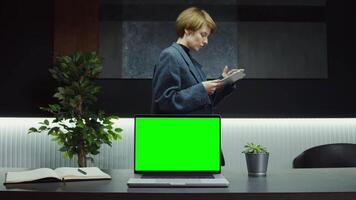 A young businesswoman uses a tablet while walking past a desktop with an open green screen laptop standing on it. Chroma key, layout, copy space. in 4k format, red komodo. video