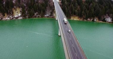 Top view of a concrete road bridge with a two-lane asphalt road with cars passing on it. drone footage of a long bridge across an emerald green river to hills covered in snowy forest. video
