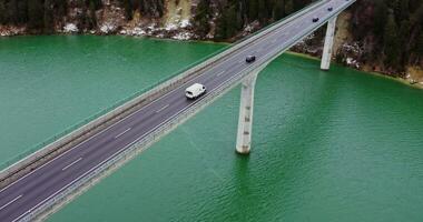 Top view of a road bridge with a two-lane asphalt road with cars passing on it. drone footage of a long bridge across an emerald green river to hills covered in snowy forest. video