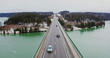 View of the bridge over the river, turning into a long road with passing cars, located among the spruce forest. filming from a drone on a winter day video