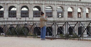 A young warmly dressed woman of European appearance takes photographs of the sights of Rome on a mobile phone, standing on an observation deck near the Coliseum. in 4k, red komodo. video