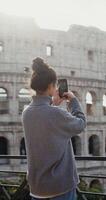 A young beautiful woman taking photos of the sights of Rome on a mobile phone, standing on an observation deck near the Coliseum. in 4k, red komodo, vertical video
