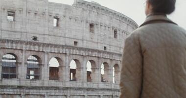 A young warmly dressed woman of European appearance comes to the observation deck in front of the Coliseum and stops looking at the amphitheater. Italy Rome. in 4k, red komodo. video