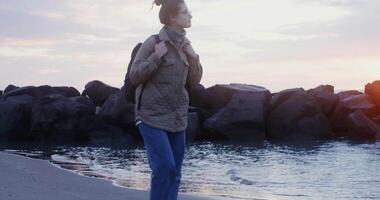 A young woman of European appearance with a large backpack on her shoulders stops at the seashore and looks into the distance while walking along the coastline at sunset. in 4k, red komodo. video