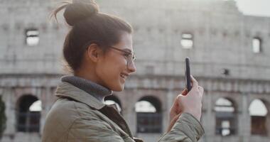 A warmly dressed young woman in glasses smiles while taking photos of the sights of Rome, standing on an observation deck near the Colosseum. shooting from the side. in 4k, red komodo. video