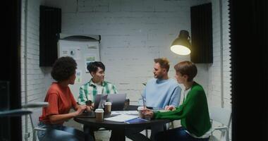 A group of young people of different nationalities are talking while sitting at a desk with laptops and work papers, discussing the business strategy of a startup video