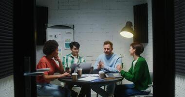 A group of young people of different nationalities are smiling while discussing the creation of a new business, sitting at the desk with many graphs and papers behind their laptops. video