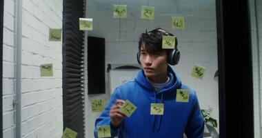 Young stylish man of asian appearance listens to music in big headphones and dances while draws arrows on the glass wall, planning, during the working day video