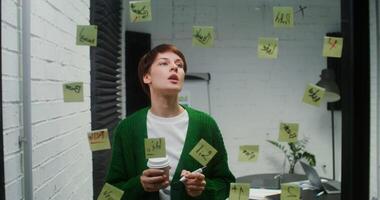 A young woman of European appearance is developing a strategy for a startup, drawing on a glass board with a marker and sticking stickers on it, working in a modern office, close-up video