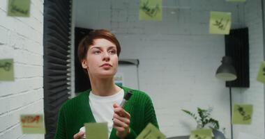 A young woman of European appearance is engaged in planning, using a glass board, stickers and markers to draw and drinks coffee from a disposable cup, working in the office alone video