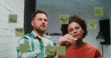 Young entrepreneurs - a man of European appearance and a African American woman are engaged in the development of a business strategy, drawing plans and charts on a glass board, in the office video