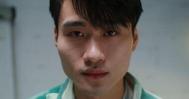 Close-up face of a young man of Asian appearance with an earring in his ear, raising his eyes from below and starting to look at the camera with a smile, blurred background video
