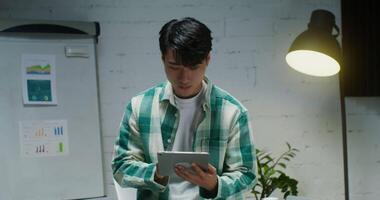 A young man of Asian appearance uses a digital tablet, while working in a stylish office and looking up from the screen looking straight into the camera with a smile video