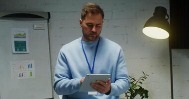 A young man of European appearance smiles while looking at the camera, interrupted from work at a digital tablet in a modern stylish office. Portrait videography of office workers video