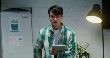 Young man of Asian appearance, dressed in casual clothes, smiling looking at camera while standing with a tablet in his hands in a modern stylish office. Portrait videography of a young businessman video
