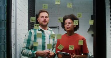 A Caucasian man and an African American woman are planning, drawing a diagram of work on a glass board with stickers and using a tablet during a working day in the office. Brainstorm, business concept video