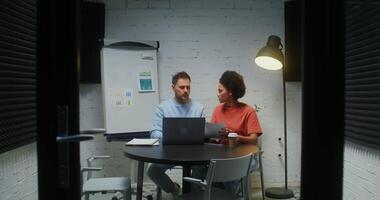 Two young colleagues - a man and a woman working in a small meeting room with a glass wall and a door, animatedly discussing work papers and using a laptop video