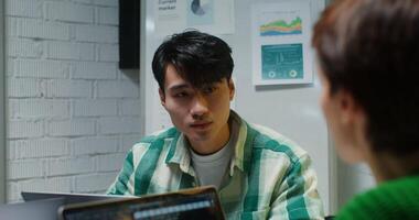Young stylish man of asian appearance, dressed in casual clothes, listens attentively to colleagues while sitting at work table with laptop, at meeting in modern office video