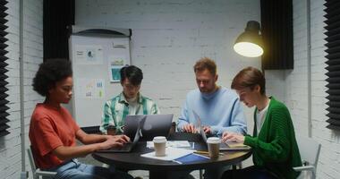 A multinational group of young entrepreneurs are working on a common project using laptops and a tablet, sitting at the same table in the meeting room and talking to each other video