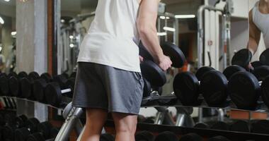 A young man of European appearance, athletic physique takes heavy dumbbells in both hands and begins to train in a modern gym, looking at himself in the mirror video