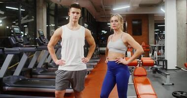 Young handsome man and woman of European appearance look directly into the camera, putting their hands on the belt, standing in a modern gym without people video