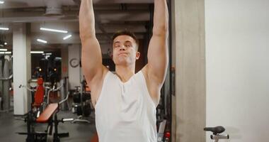 Front view of a young athletic man of European appearance doing pull-ups on the simulator in a modern gym, close-up video