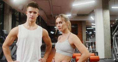 Young man and woman of European appearance in sportswear, looking directly at the camera, standing in the middle of a well-equipped gym, portraits, close-up video