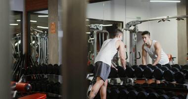 A young man of European appearance, athletic physique starts workshop for biceps in a modern gym, standing in front of a mirror and choosing dumbbells video