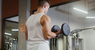 A young man athletic physique trains biceps, lifting a heavy dumbbell with one hand and looking at himself in the mirror, standing in the gym, filming from behind, man's face is not visible video