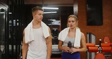 Young handsome man and woman of European appearance with towels around their necks are talking, while walking in a modern gym and choosing which exercise machine to train on video