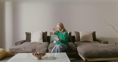 Elderly gray-haired woman of European appearance, dressed in casual clothes, smiling while talking on a call on a tablet while sitting on a sofa in a modern home interior video