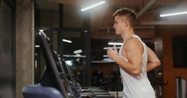 Young man of european appearance athletic physique trains on the running on a treadmill, exercising in a modern gym alone, close-up, side view video