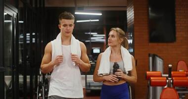 Young man and woman of European appearance with towels around their necks are talking while walking in a modern gym, intending to work out on a treadmill video
