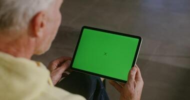 An elderly gray-haired man dressed in casual clothes chat on a tablet with a green screen in his hands. Filming from behind, the man's face is not visible, chroma key, copy space video