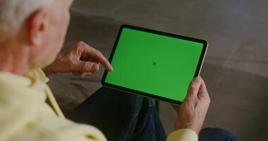 An elderly gray-haired man dressed in casual clothes uses a tablet, scrolling the screen with a green screen, filming from behind, the man's face is not visible, chroma key, copy space video