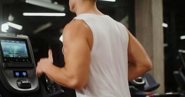 A young man of European appearance, athletic build, runs on a treadmill, looking intently at the digital display. Close-up of the torso and strong arms in half-turn, close-up video