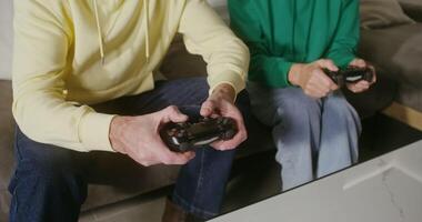 Old man and woman, dressed in casual clothes, are enthusiastically playing an online game, sitting on the couch at home and using joysticks, actively communicating and trying to beat each other video