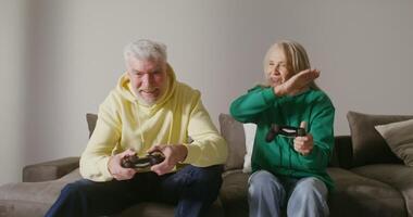 An elderly man and woman are enthusiastically playing an online game, sitting on the couch and using joysticks, actively communicating and trying to beat each other video