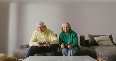 An elderly man and woman are playing a computer game, sitting on the couch with joysticks in their hands, laughing and talking cheerfully, without stopping from the game, in a stylish home environment video