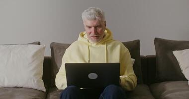 An elderly gray-haired man of European appearance smiling starts a call, waving hello while sitting on a sofa in a modern home interior video