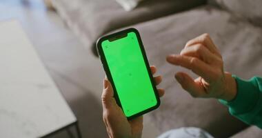 An elderly woman uses a mobile phone while scrolling the screen while sitting on the couch. filming from behind, female hands with green screen phone, close-up, chroma key, copy space video