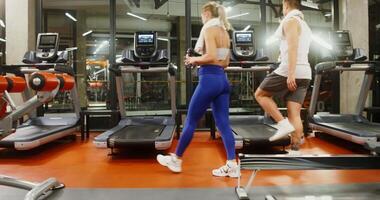 A young man and woman of European appearance walk up to a modern treadmill and start the workout together. Rear view of young couple running on treadmill in gym video