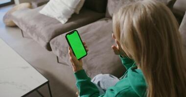 Elderly woman waves hello starting a call on a green screen mobile phone she is holding in front of her, filming from behind, chroma key, copy space video